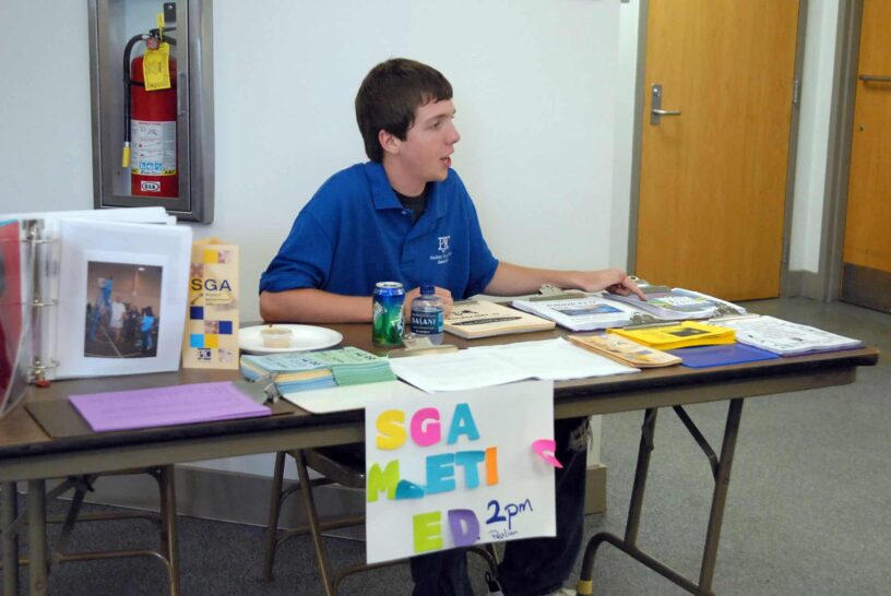 Student government member sitting at table with flyers and sign at eCorsair event.