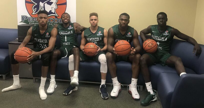 Pensacola State College basketball players sitting with basketballs in locker room.