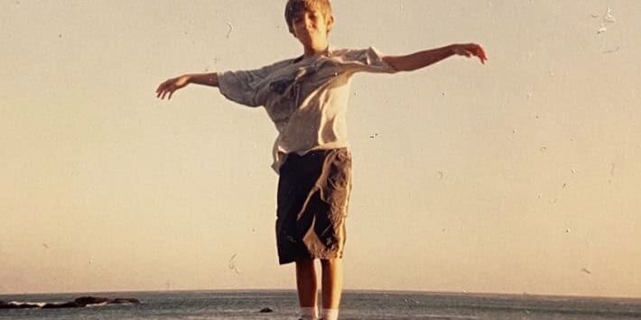Young boy standing on a rock with arms outstretched by the ocean at sunset, symbolizing reflection a.