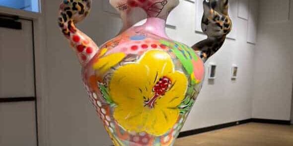 Colorful ceramic vase with hand-shaped handles and floral design, displayed in an art gallery.
